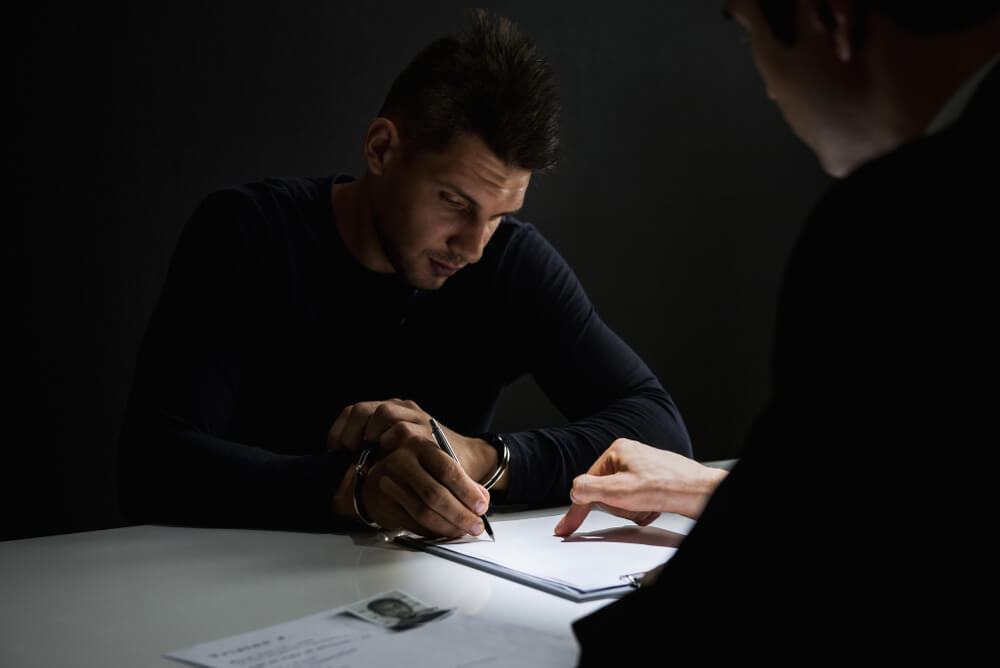 Criminal man with handcuffs signing document in interrogation room.